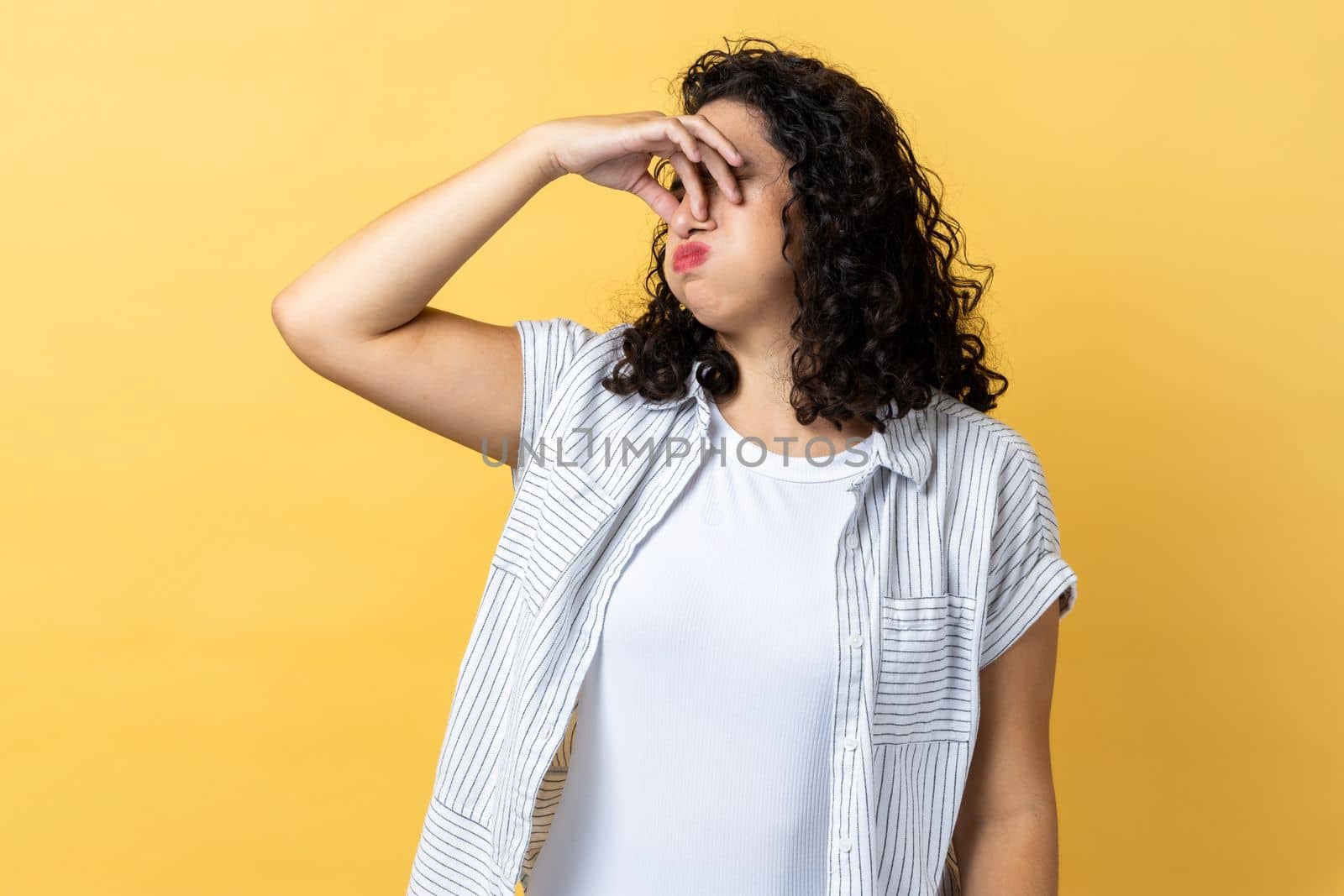 Awful smell. Woman with dark wavy hair pinching nose, stop breathing bad odor, disgusted by smell of farting, her grimace expressing repulsion. Indoor studio shot isolated on yellow background.
