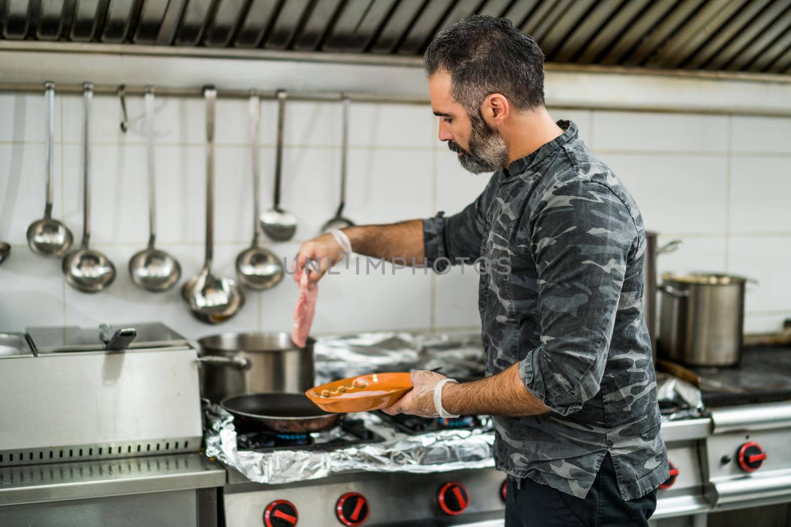 Professional cook is preparing meal in restaurant's kitchen. He is frying meat.