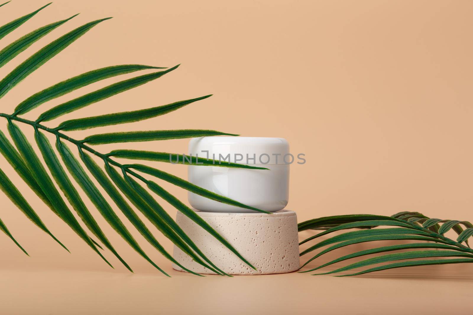 White cosmetic jar on podium against pastel beige background decorated with palm leaves. Concept of organic eco skin care products with nourishing, moisturizing effect