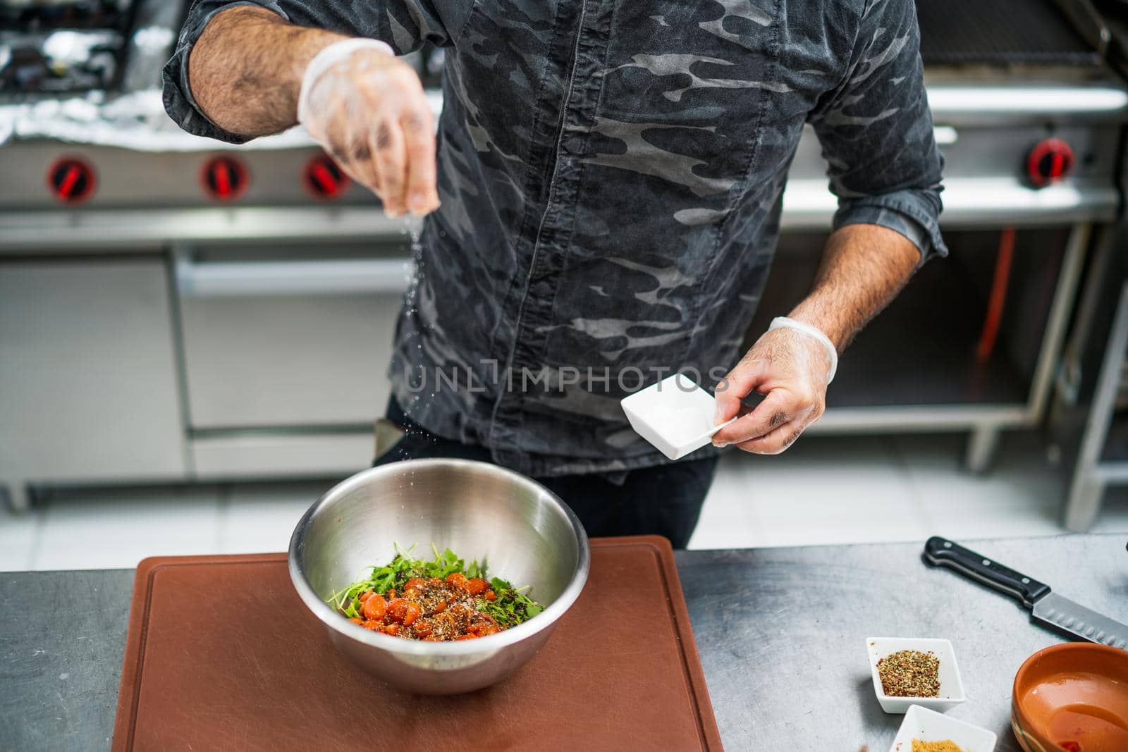 Professional cook is preparing meal in restaurant's kitchen. He is salting salad.