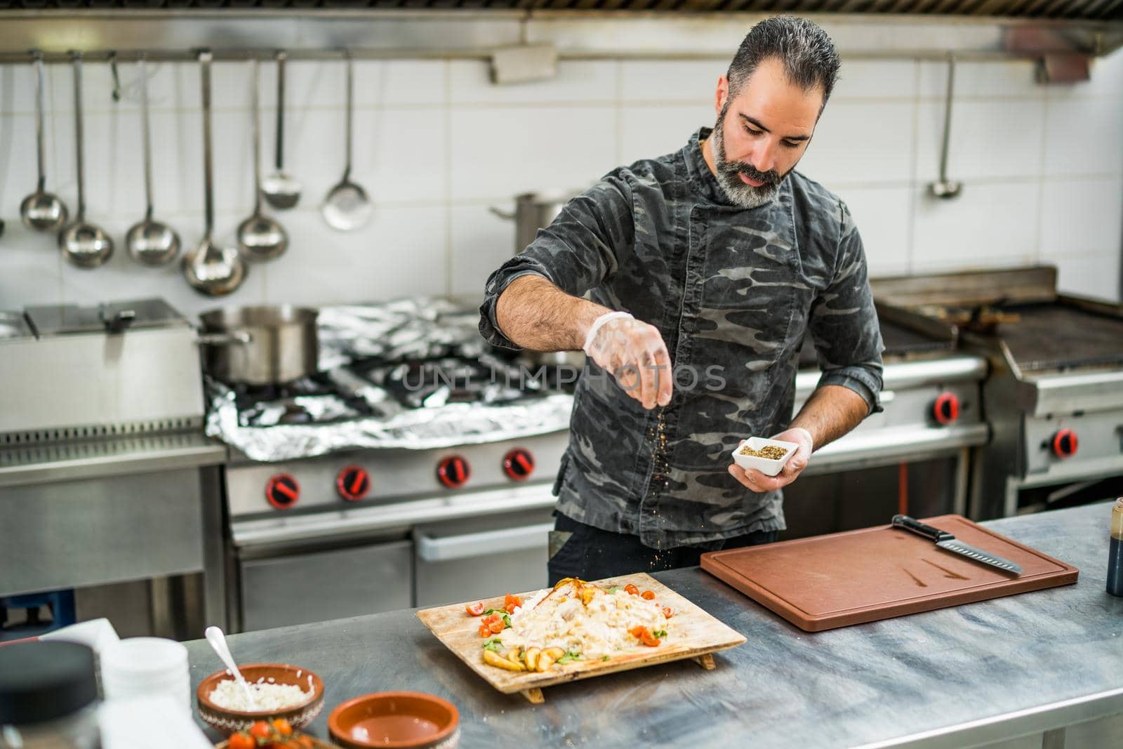 Professional cook is preparing meal in restaurant's kitchen. He is inserting spices in meal after cooking.