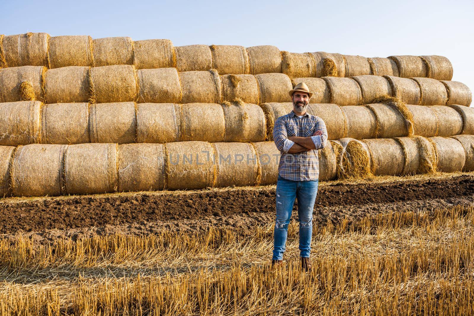 Happy farmer is standing beside bales of hay. He is satisfied because of successful harvesting.