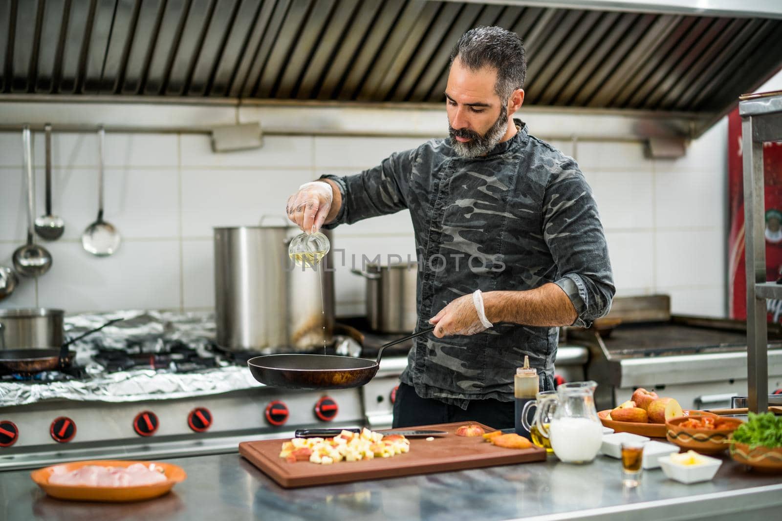 Professional cook is preparing meal in restaurant's kitchen. He is oiling pan before frying meat.