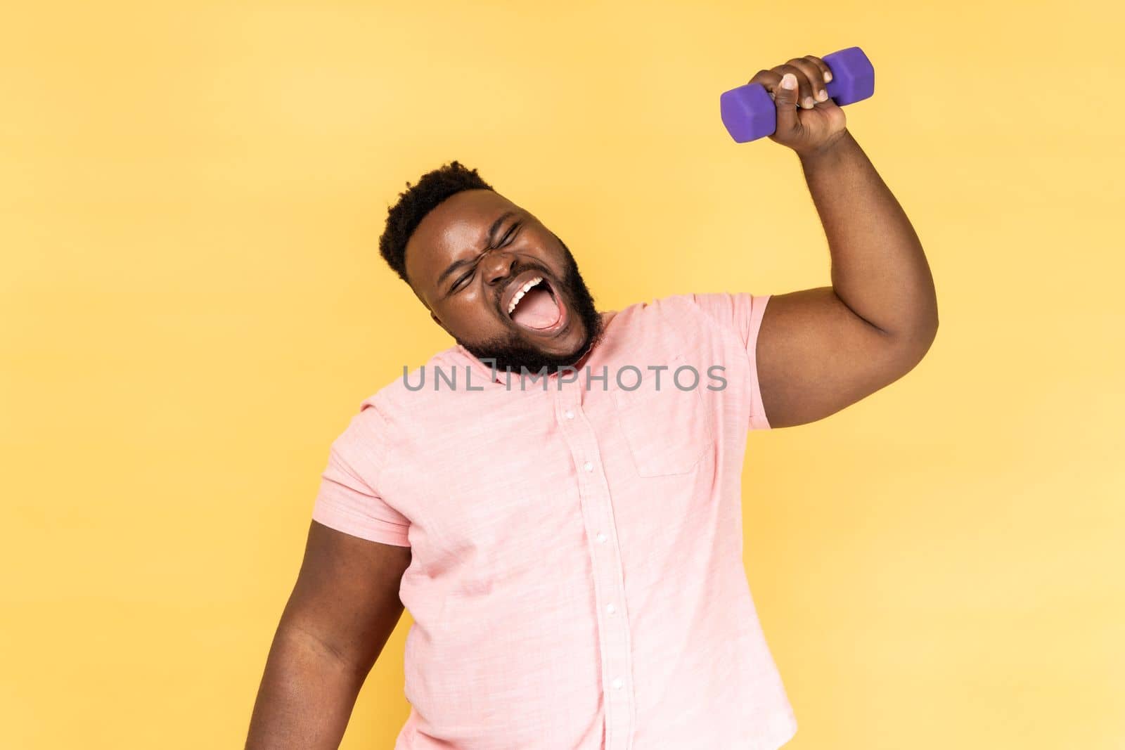 Portrait of funny positive bearded man wearing pink shirt standing and raised arm with dumbbell, making effort raising heavy sport equipment. Indoor studio shot isolated on yellow background.