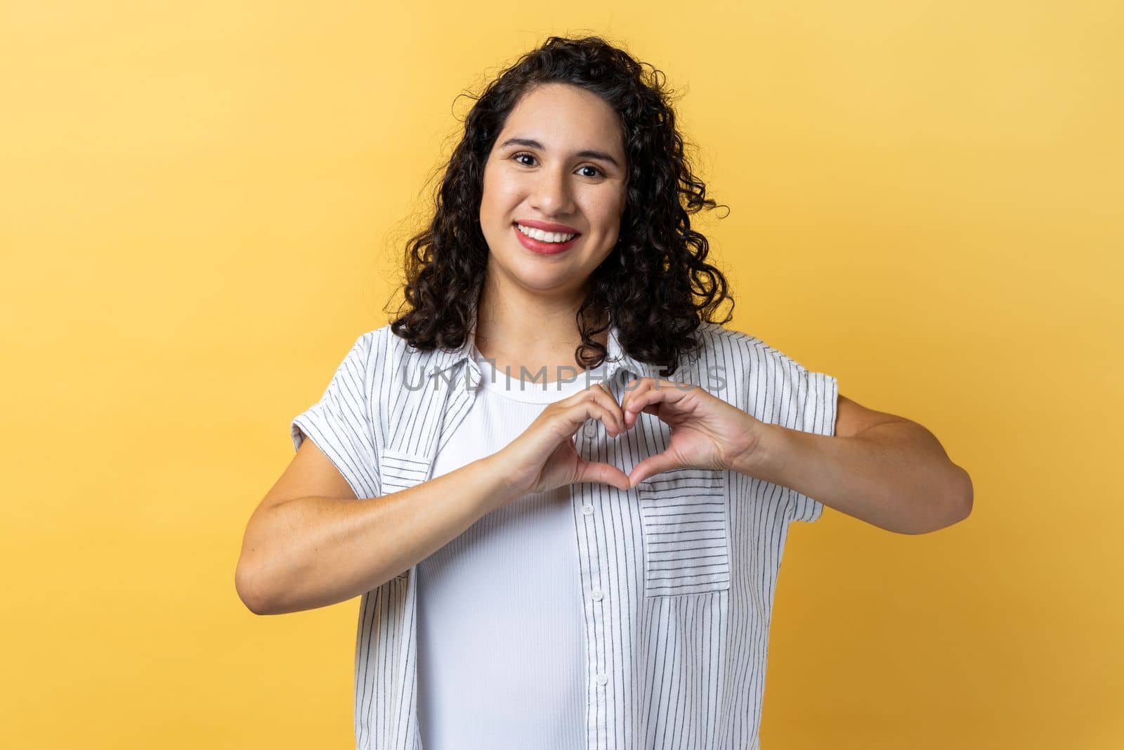 Portrait of woman with dark wavy hair holding hands in shape of heart showing romantic gesture, love confession, valentines day celebration. Indoor studio shot isolated on yellow background.