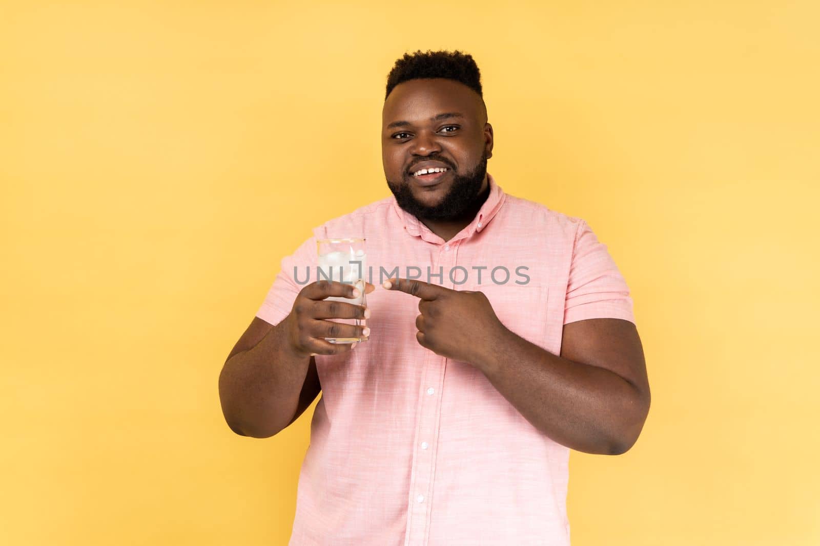 Portrait of smiling satisfied bearded man wearing pink shirt holding glass of water with ice, pointing at cold beverage, looking at camera. Indoor studio shot isolated on yellow background.