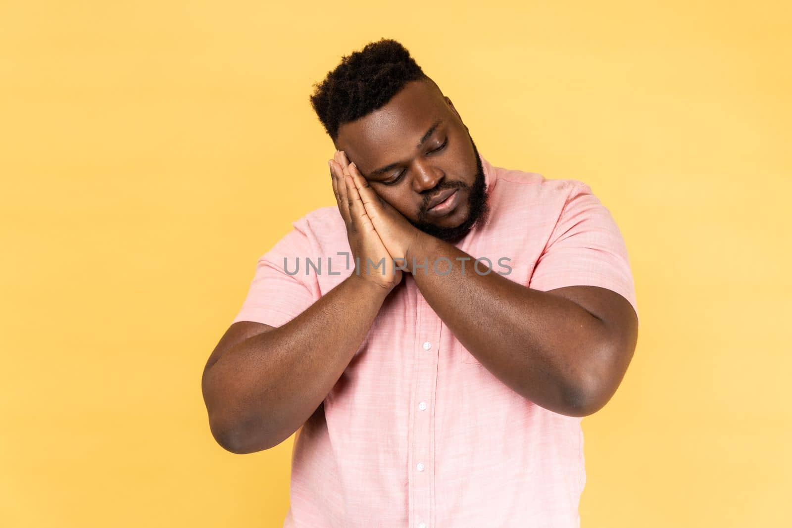 I want to sleep. Portrait of man wearing pink shirt with closed eyes standing with arms on her cheeks like pillow and sleep, having nap. Indoor studio shot isolated on yellow background.