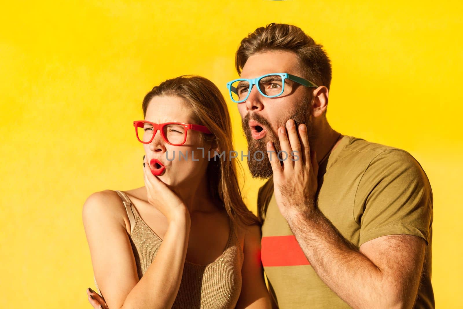 Portrait of amazed young adult couple in colorful glasses looking away with astonished facial expression, being shocked and surprised. Indoor studio shot isolated on yellow background.