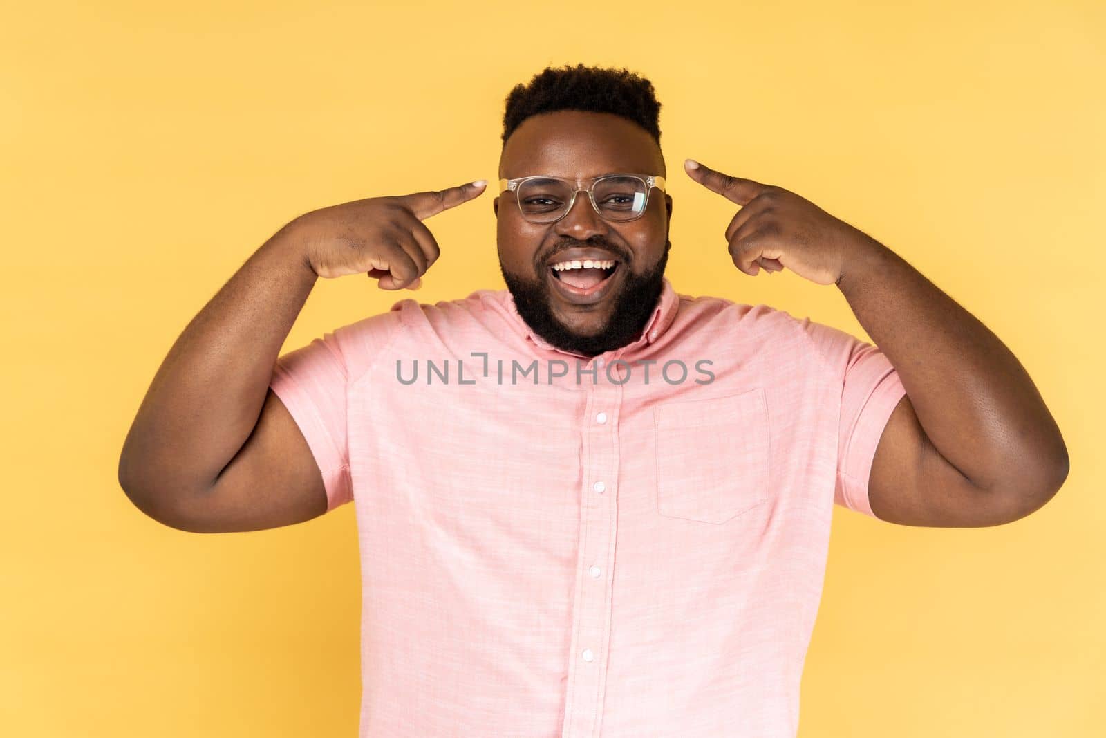 Portrait of delighted satisfied man wearing pink shirt standing, pointing at his new optical spectacles, being happy to see better, smiling happily. Indoor studio shot isolated on yellow background.