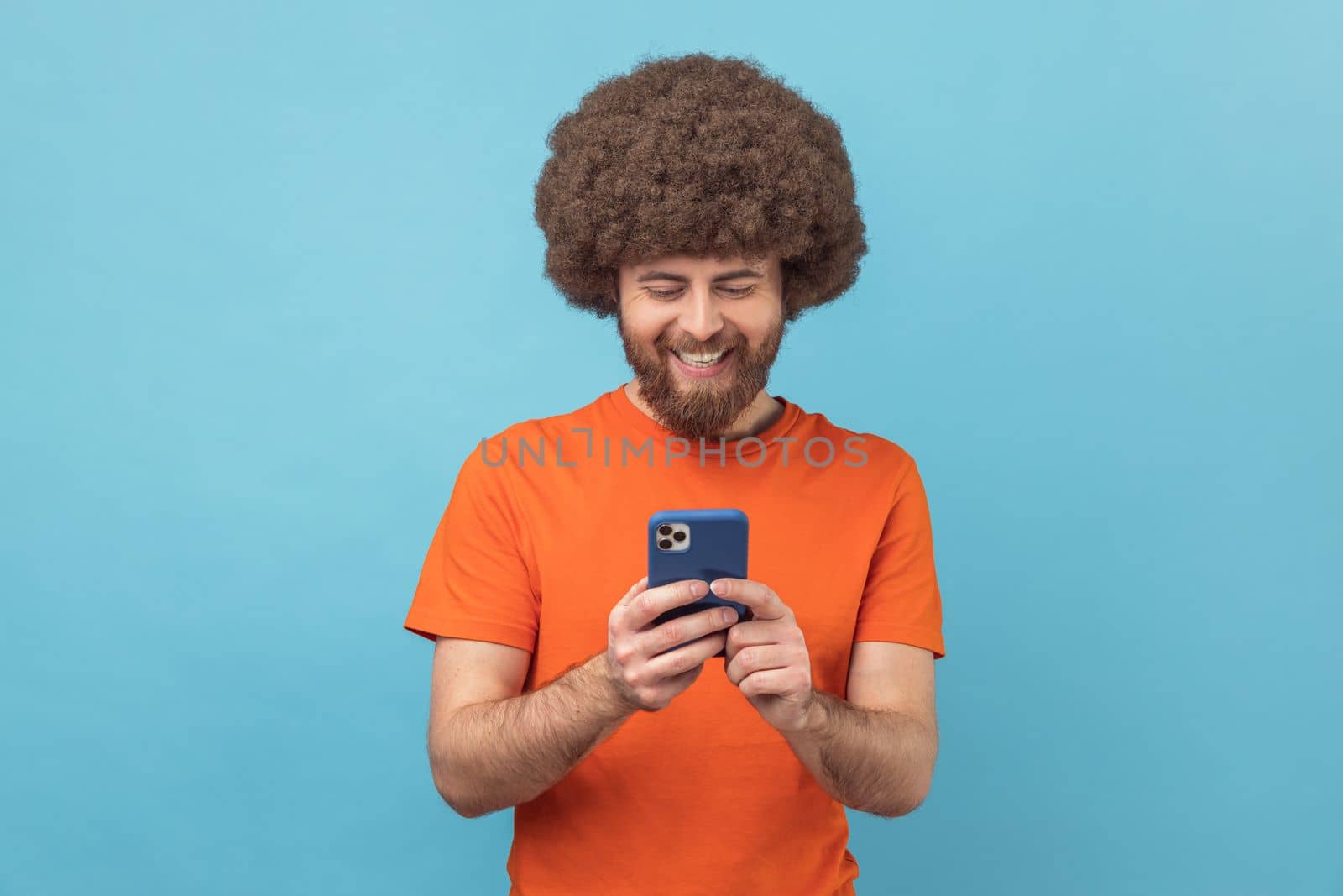 Portrait of positive man blogger with Afro hairstyle wearing orange T-shirt standing, using smartphone and watching video with happy smiling face. Indoor studio shot isolated on blue background.