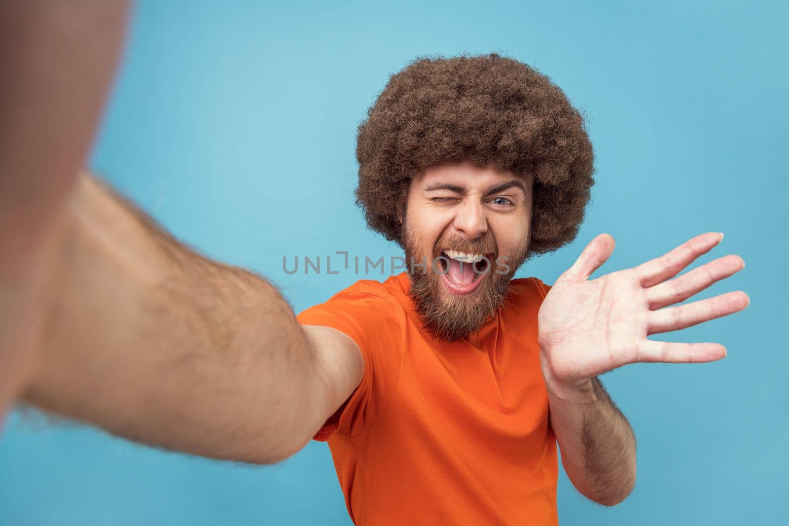 Portrait of excited man with Afro hairstyle wearing orange T-shirt having livestream, talking with followers, waving hand, greeting, POV. Indoor studio shot isolated on blue background.