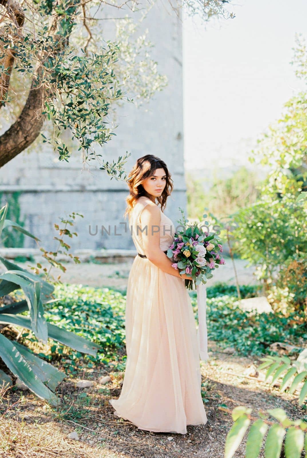 Bride with a bouquet stands half-turned near a green tree. High quality photo