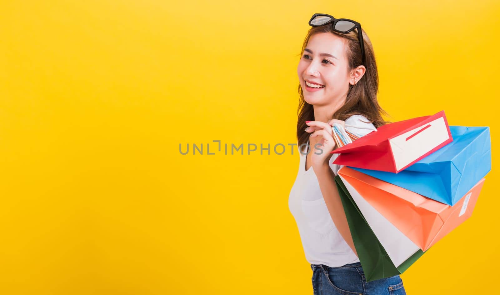 Asian Thai portrait happy beautiful cute young woman smiling stand with sunglasses excited holding shopping bags multi color looking side, studio shot isolated yellow background with copy space