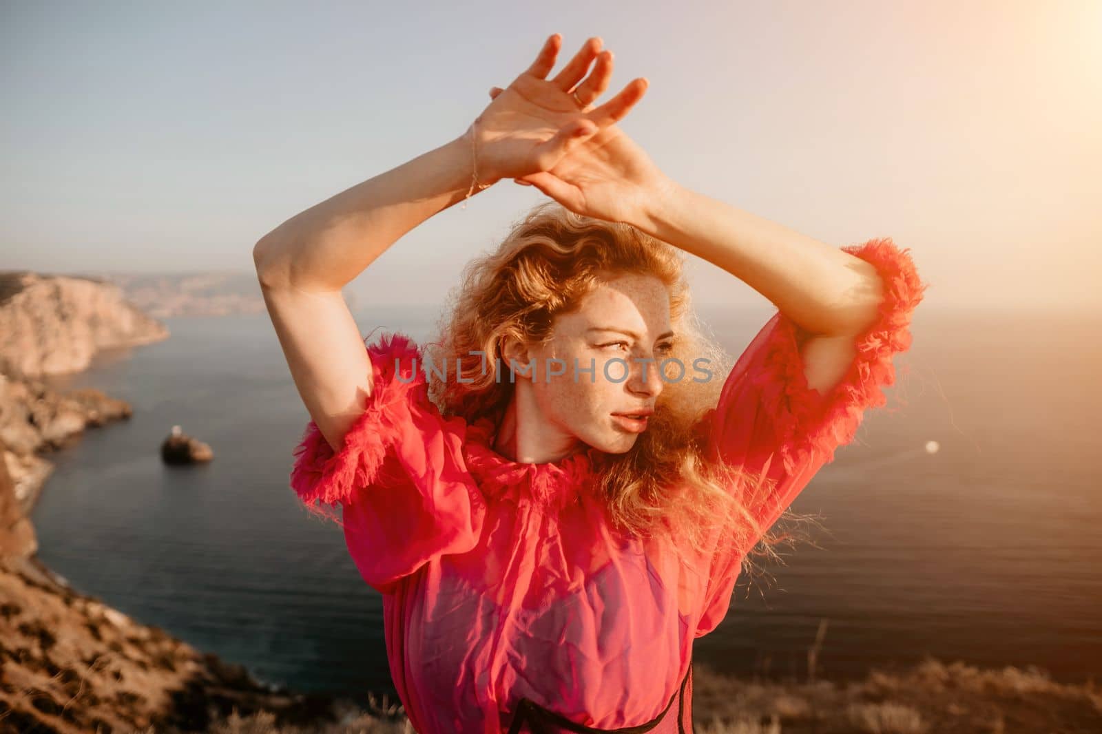 Close up shot of beautiful young caucasian woman with curly blond hair and freckles looking at camera and smiling. Cute woman portrait in a pink long dress posing on a volcanic rock high above the sea