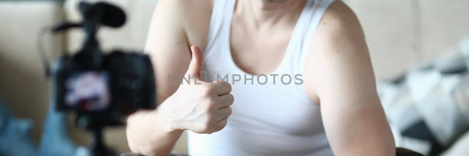 Athlete man holds thumbs up and raises dumbbells sitting on ball at home in front of camera. Recommendations for home fitness and sports training remotely