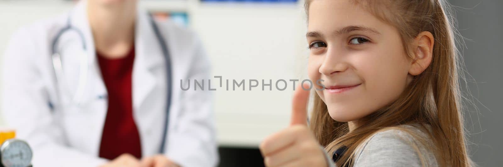 Beautiful little girl shows thumbs up. and doctor sits in background. Health insurance and examination of children