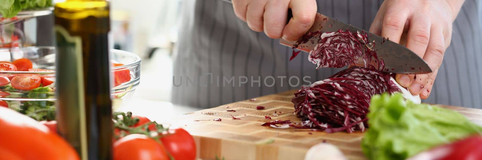 Male hands are chopping red cabbage on cutting board surrounded by various fresh vegetables on table. Closeup of male hands slicing fresh vegetables on cutting board