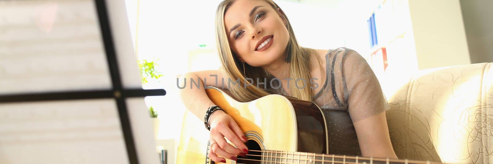 Smiling blond woman playing guitar and looking through notes. Talented musician and music hobby concept