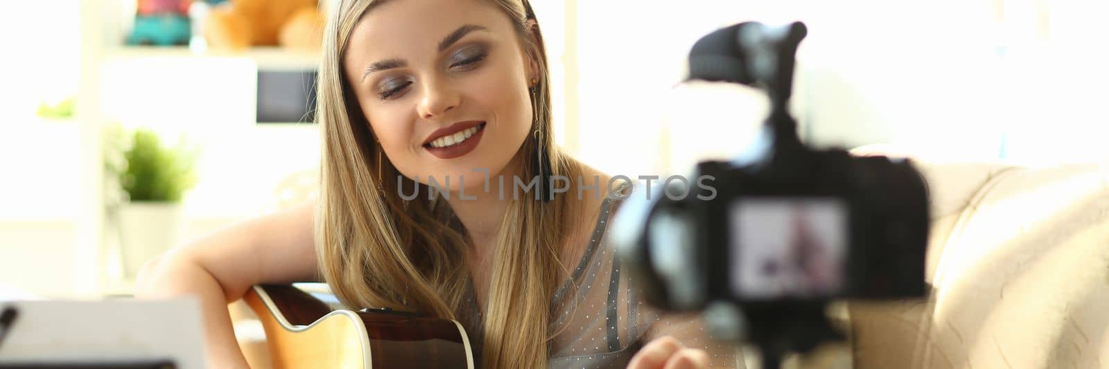 Young woman plays acoustic guitar and records video clips for social networks. Amateur musician creates content at home using modern camera concept