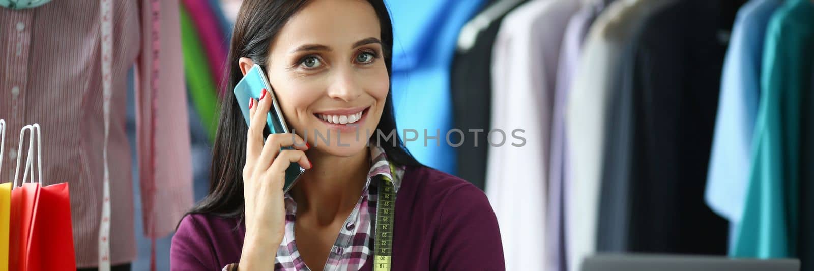 Attractive experienced seamstress needlewoman at workplace in atelier calling to client. Fashion stylist designer discussing on smartphone individual design of fabric clothing at workplace