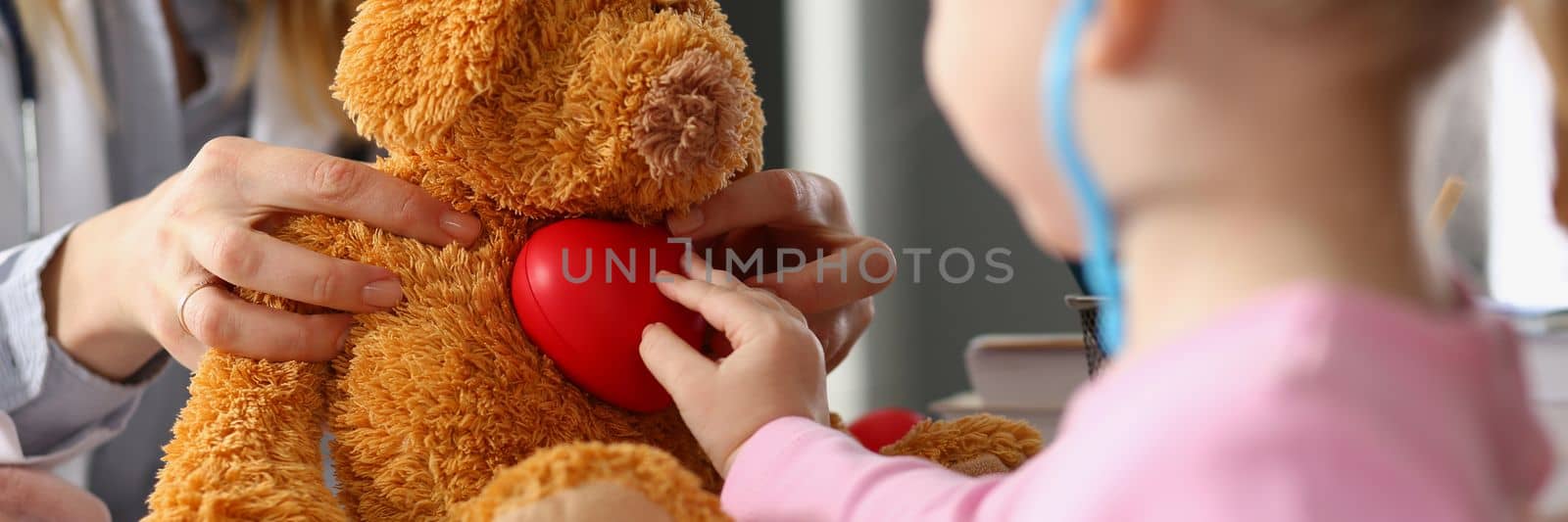 Little child girl plays with plush toy at doctor appointment. Child listens to soft toy with stethoscope and holds heart. Treatment of cardiac diseases in children