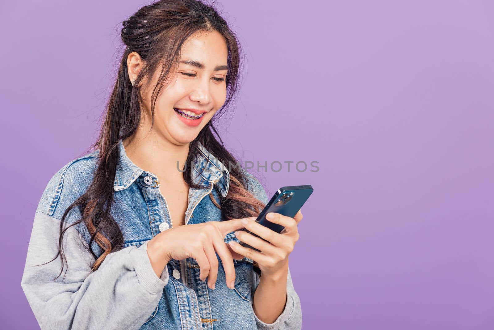 Portrait of Asian beautiful young woman confident wear denim jeans shirt hold smartphone typing smartphone, Happy lifestyle female write message look phone, studio shot isolated on purple background