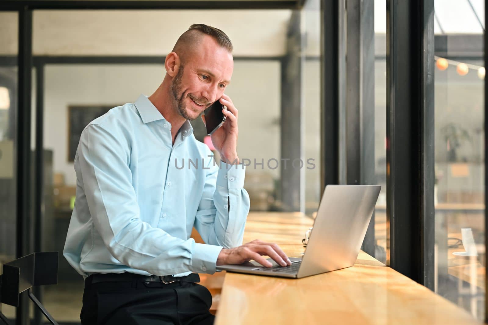 Smiling caucasian businessman talking on mobile phone and checking information on laptop computer at coffee shop by prathanchorruangsak