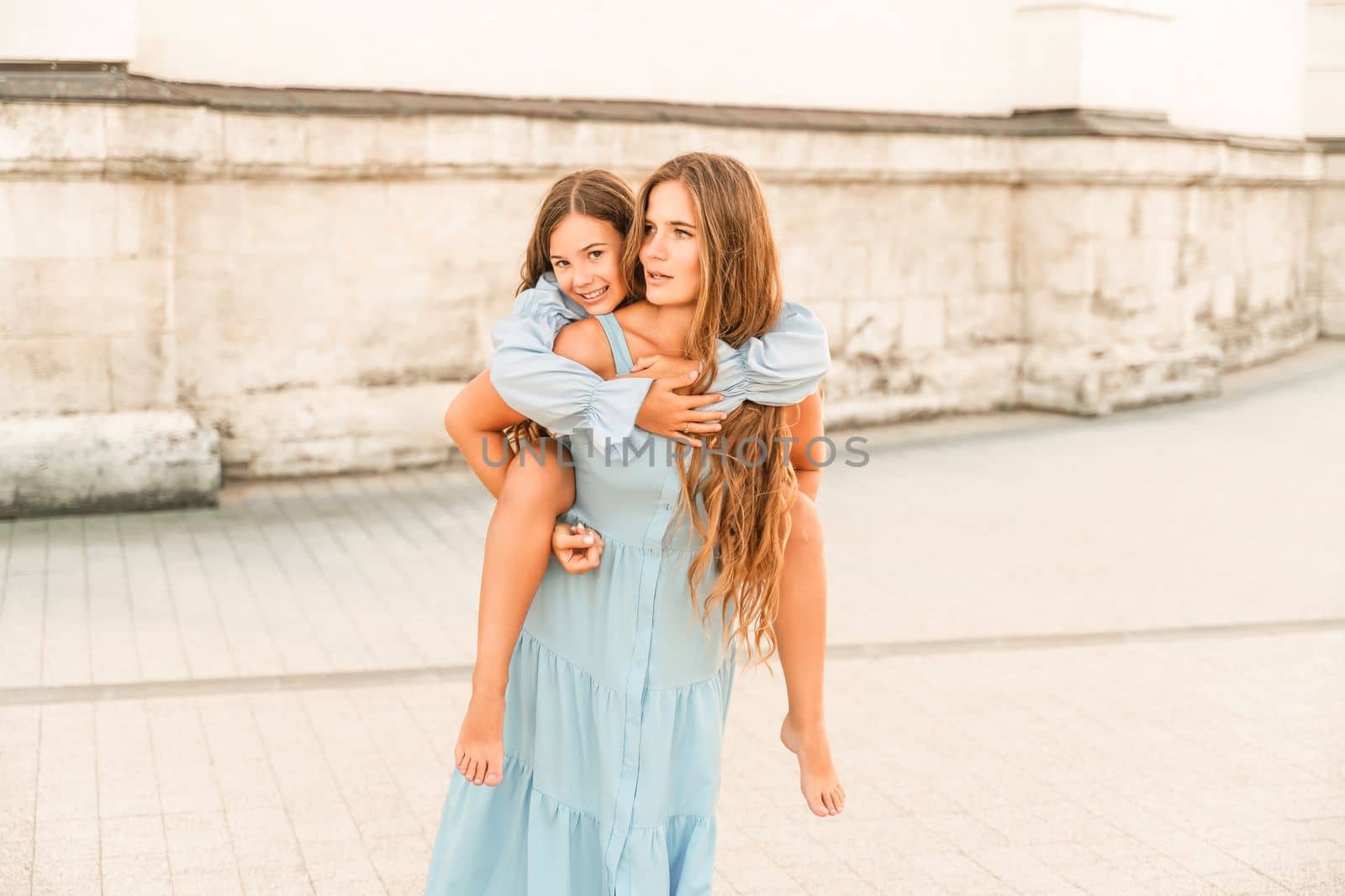 Mother of the daughter walks playing. Mother holds the girl on her back, holding her legs, and her daughter hugs her by the shoulders. Dressed in blue dresses. by Matiunina