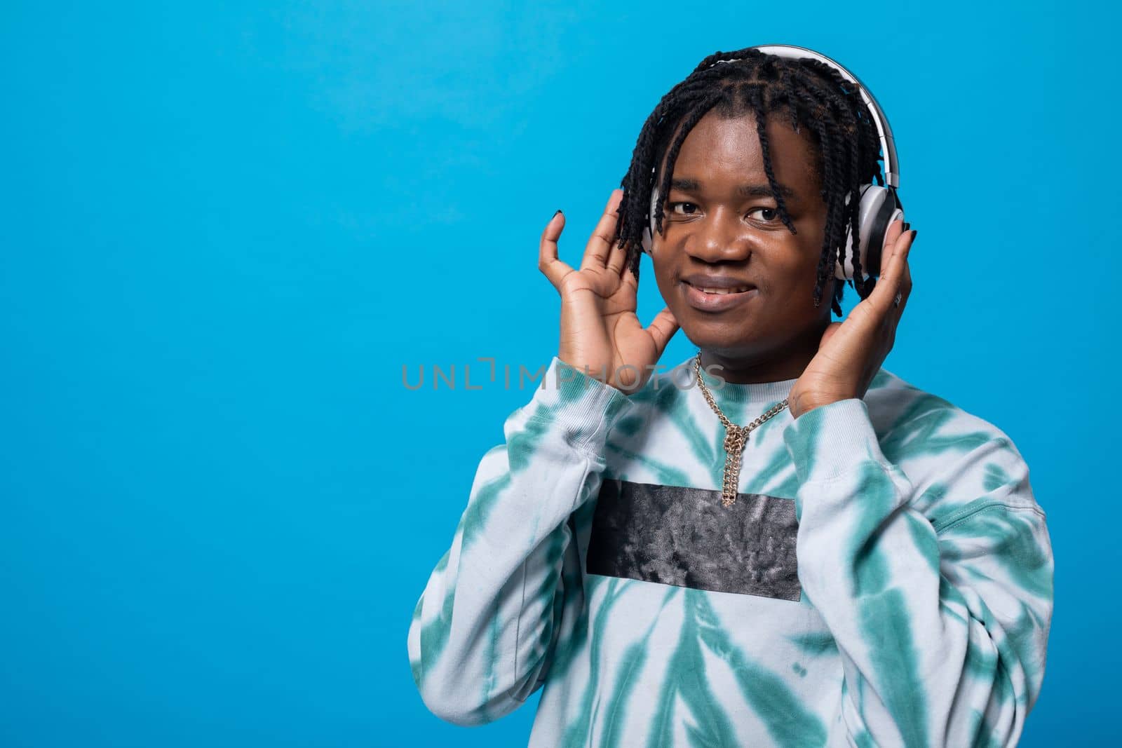 Against a blue solid background, a dark-skinned man has headphones on his ears and looks straight into the camera. A smiling boy in dreadlocks.
