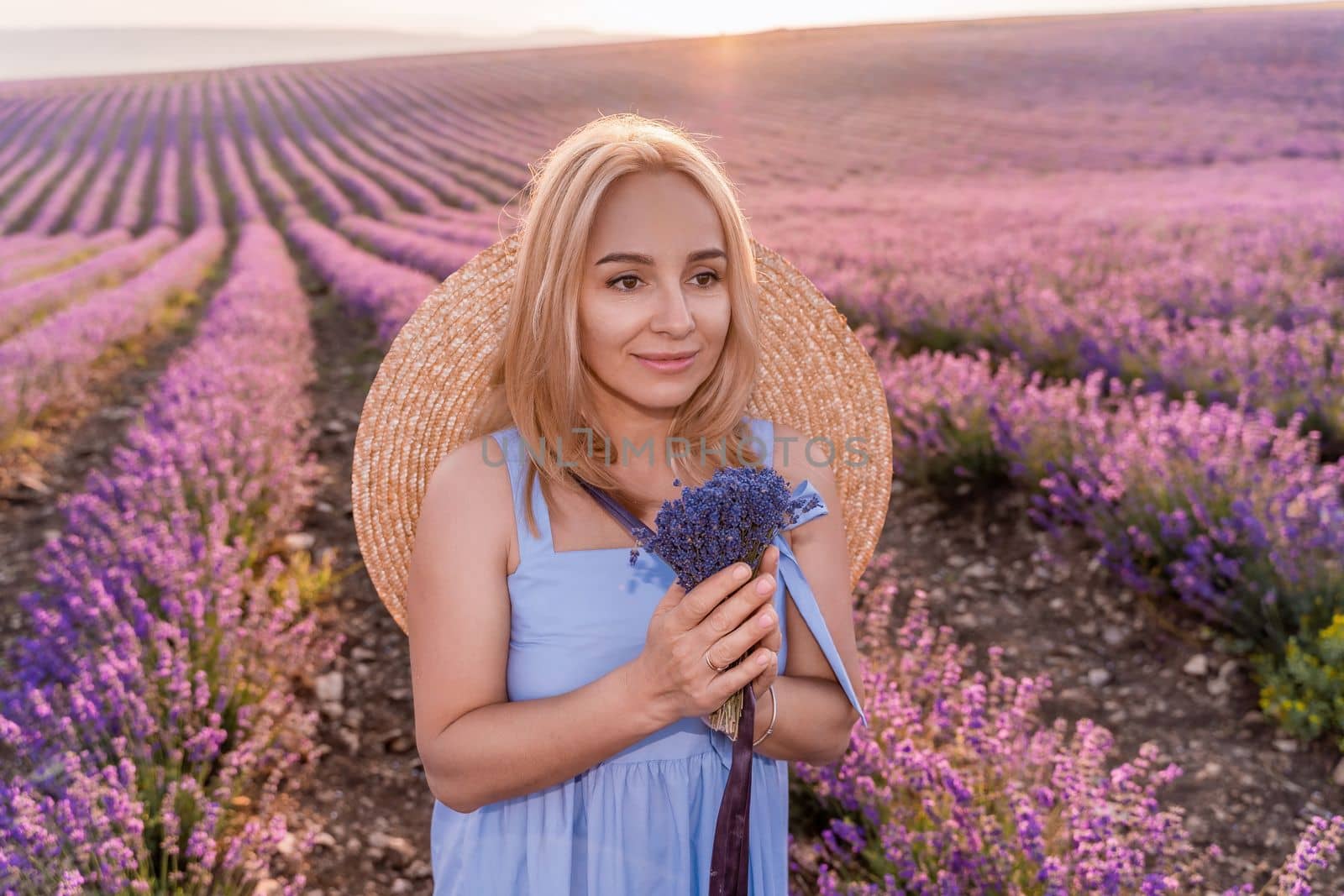 Woman lavender field sunset. Romantic woman walks through the lavender fields. illuminated by sunset sunlight. She is wearing a blue dress with a hat