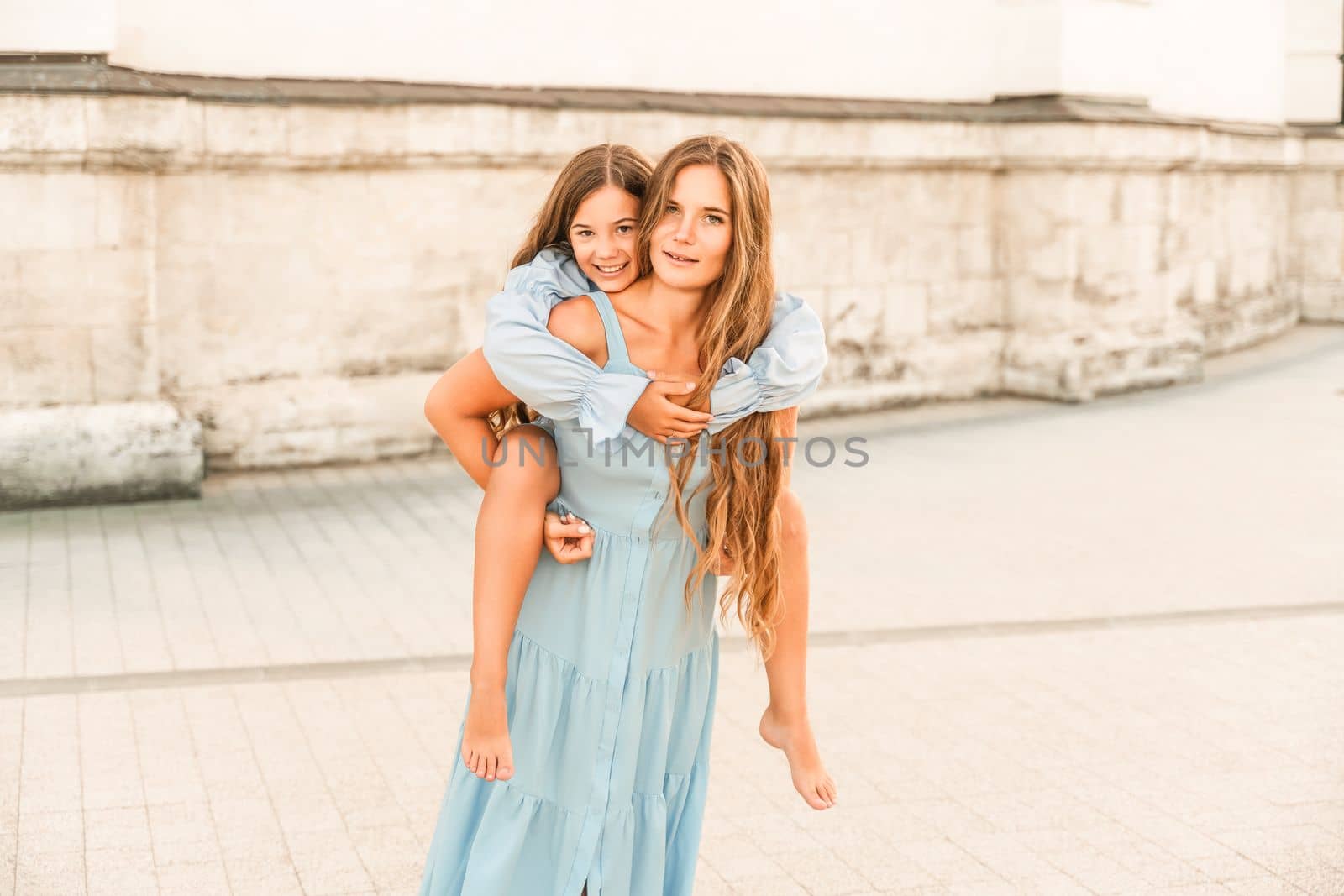 Mother of the daughter walks playing. Mother holds the girl on her back, holding her legs, and her daughter hugs her by the shoulders. Dressed in blue dresses. by Matiunina