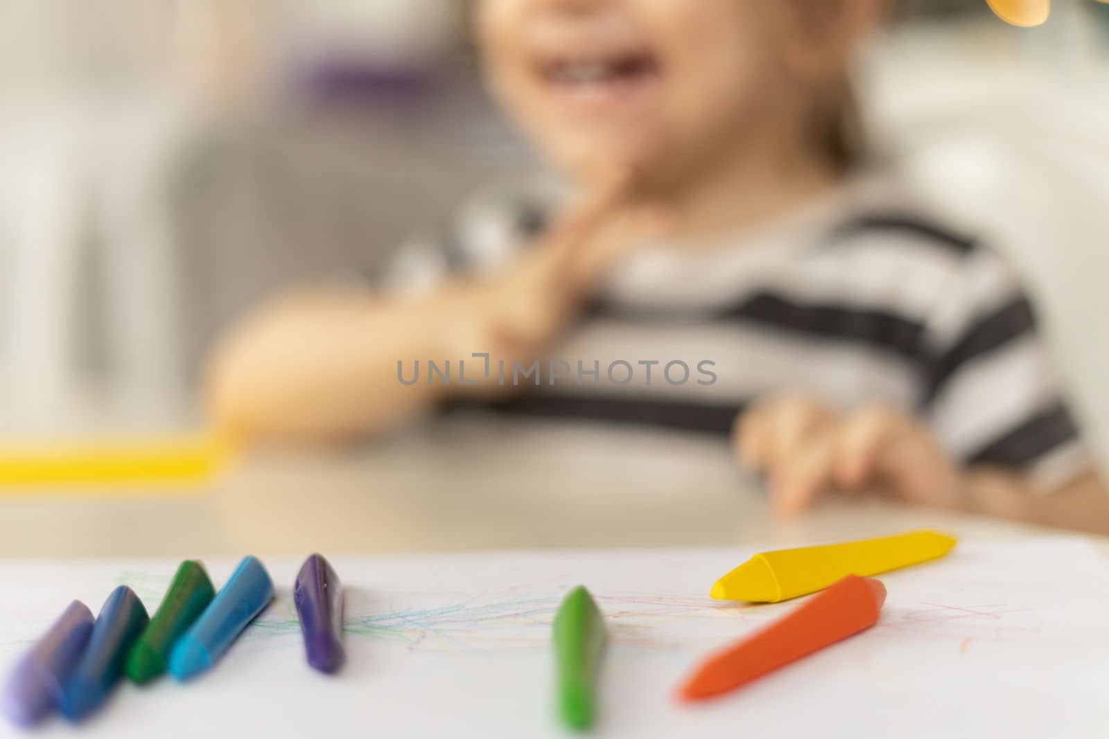 Selective focus on colorful crayons. Smiling caucasian baby girl sitting at the table at home drawing abstract picture.