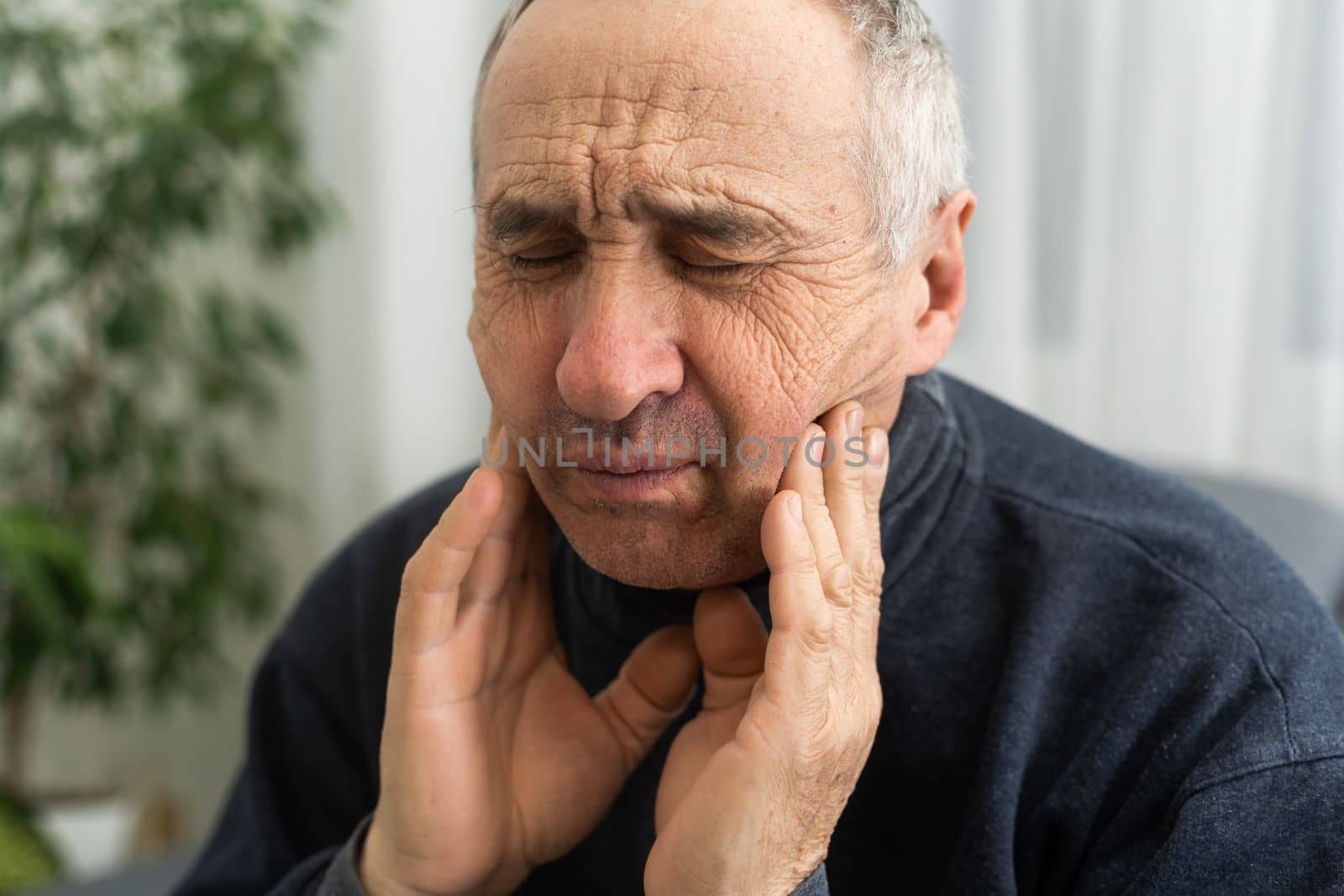 Old man with toothache. Elderly senior man has toothache. Unhappy man face in tooth pain sitting on sofa at home, feel sick unwell. Sad aged man hand holding his chin. Adult suffering toothache.