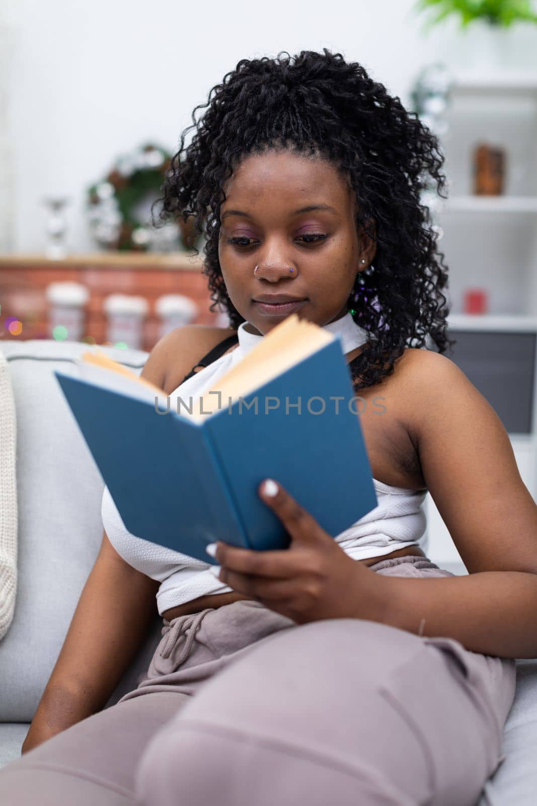 A college student studies while reading a book. A dark-skinned girl with black and curly hair sits on a sofa. Christmas decoration.