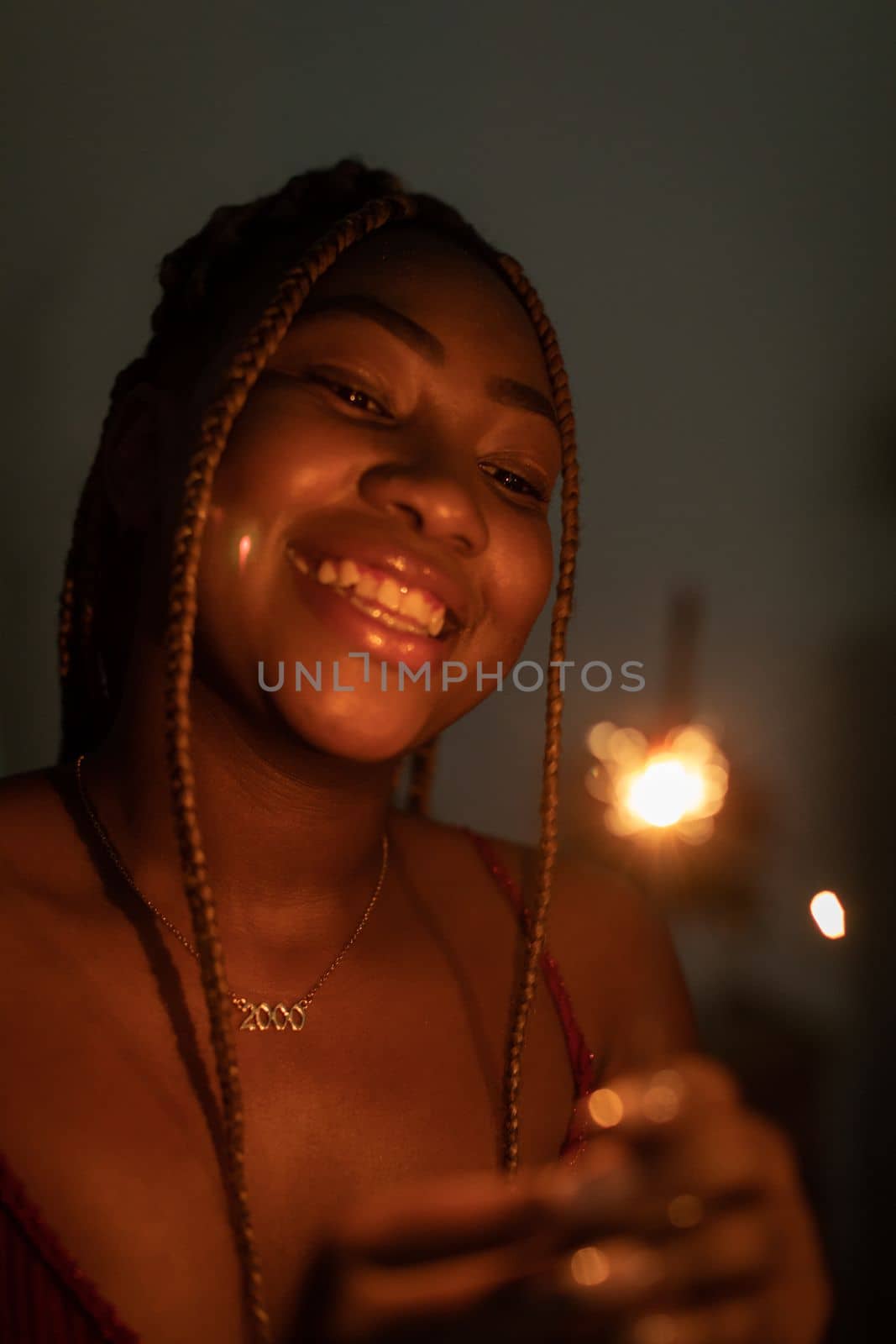 Against a dark background, a dark-skinned girl stands and smiles while holding burning cold fires in her hand. Happy New Year celebration.