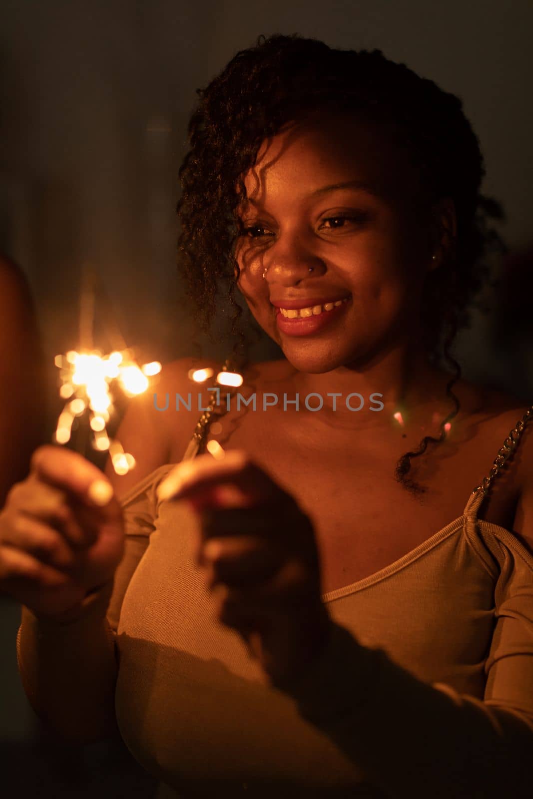 Against a dark background, a dark-skinned girl stands and smiles while holding burning cold fires in her hand. Happy New Year celebration.