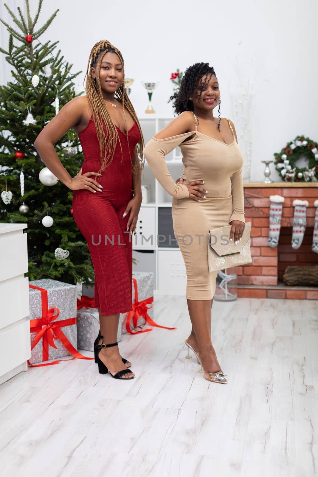 Happy and elegantly dressed two dark-skinned girls wait for invited guests at a decorated Christmas tree. Room decorated for Christmas.