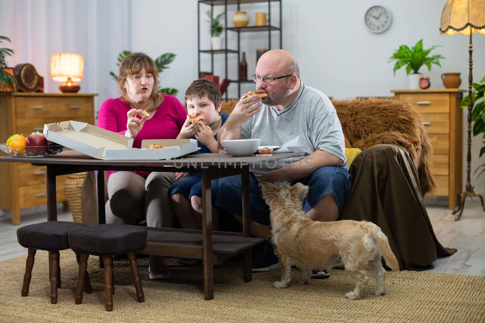 A shaggy dog watches when he too gets his slice. Mother, Father and son help themselves to hot and fragrant slices of pizza from a box.