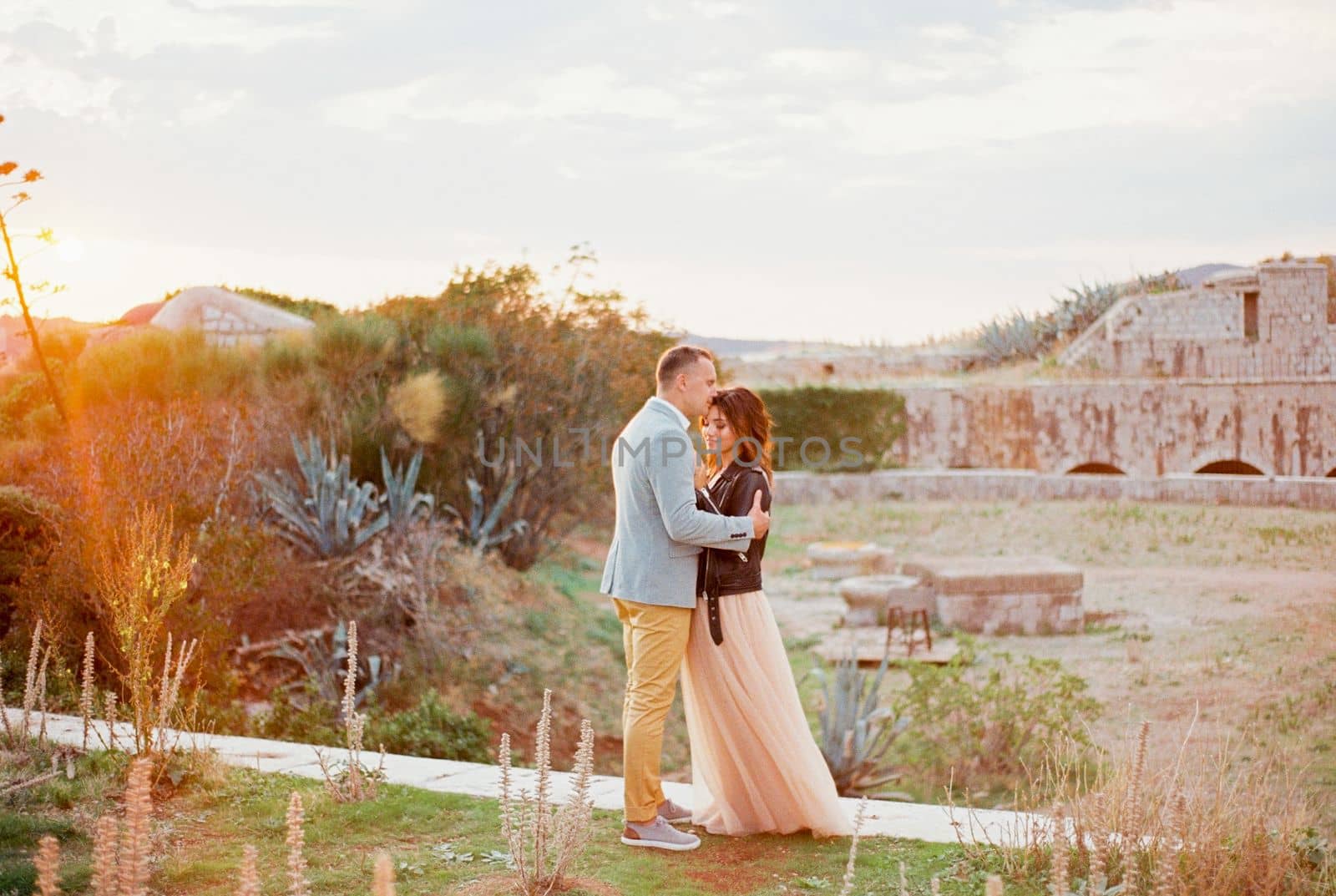 Groom hugs and kisses bride on the forehead standing on a stone path. High quality photo