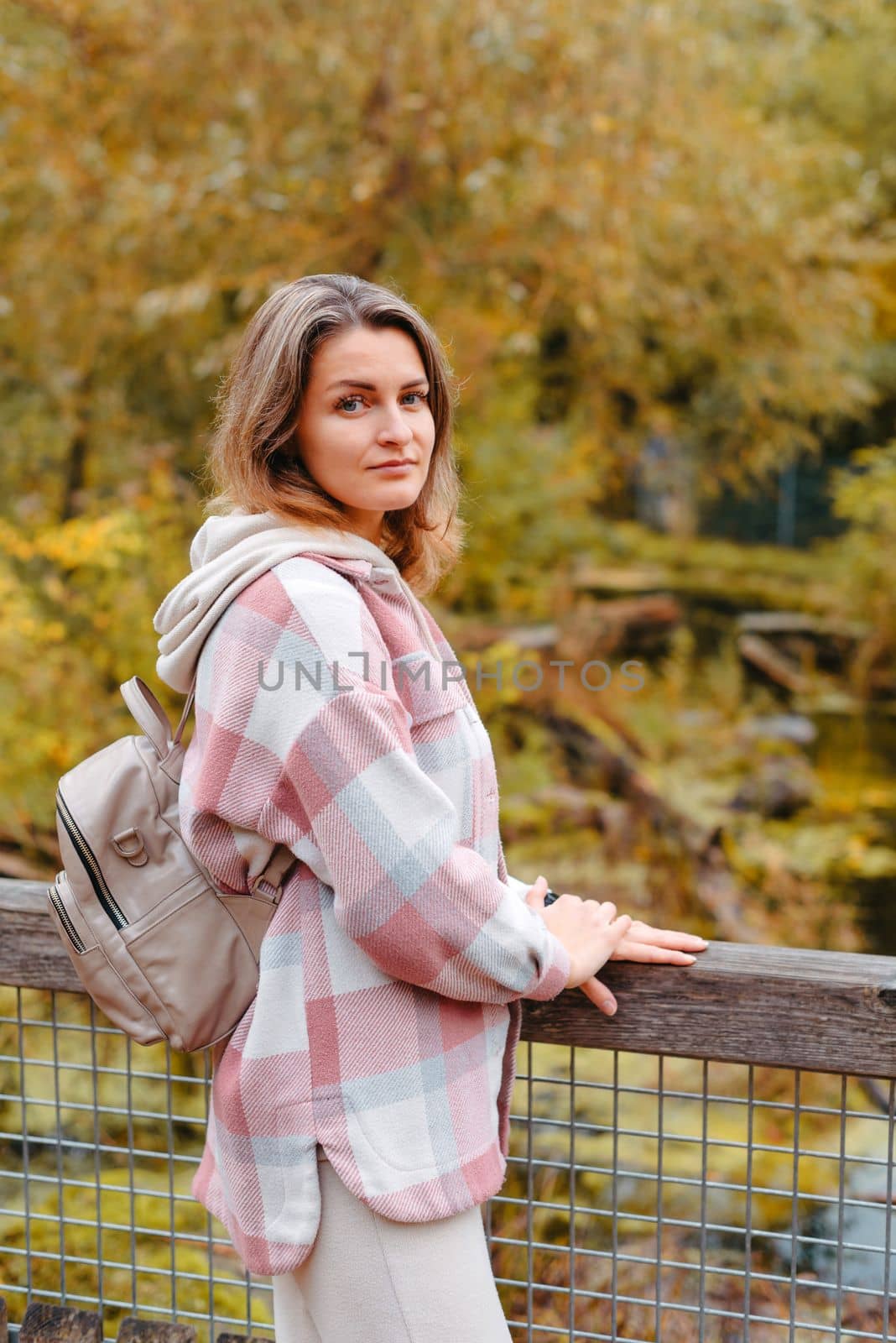 Portrait Of Cute Young Woman In Casual Wear In Autumn, Standing On Bridge Against Background Of An Autumn Park And River. Pretty Female Walking In Park In Golden Fall. Copy Space. Smiling Girl In The Park Standing On Wooden Bridge And Looking At The Camera In Autumn Season by Andrii_Ko