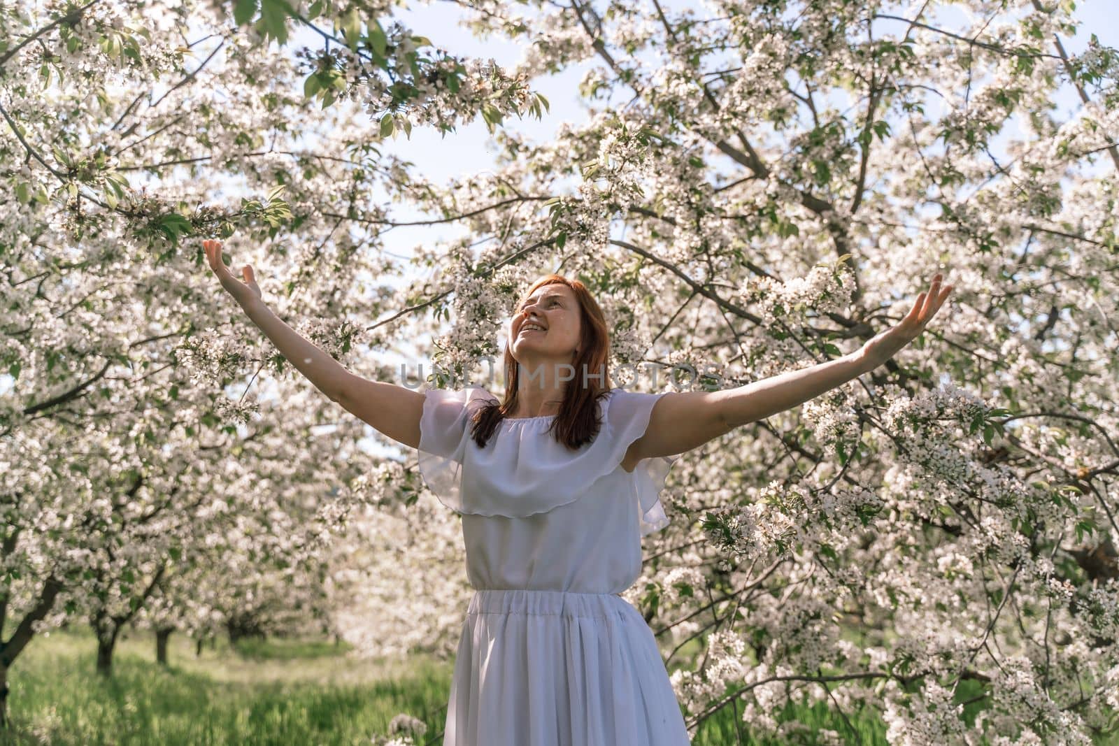 Fantasy woman in long white elegant fashion long dress walks in green spring blossom cherry garden. Happy cheerful girl princess bride. Skirt fabric flies flowing waving in wind motion. by Matiunina