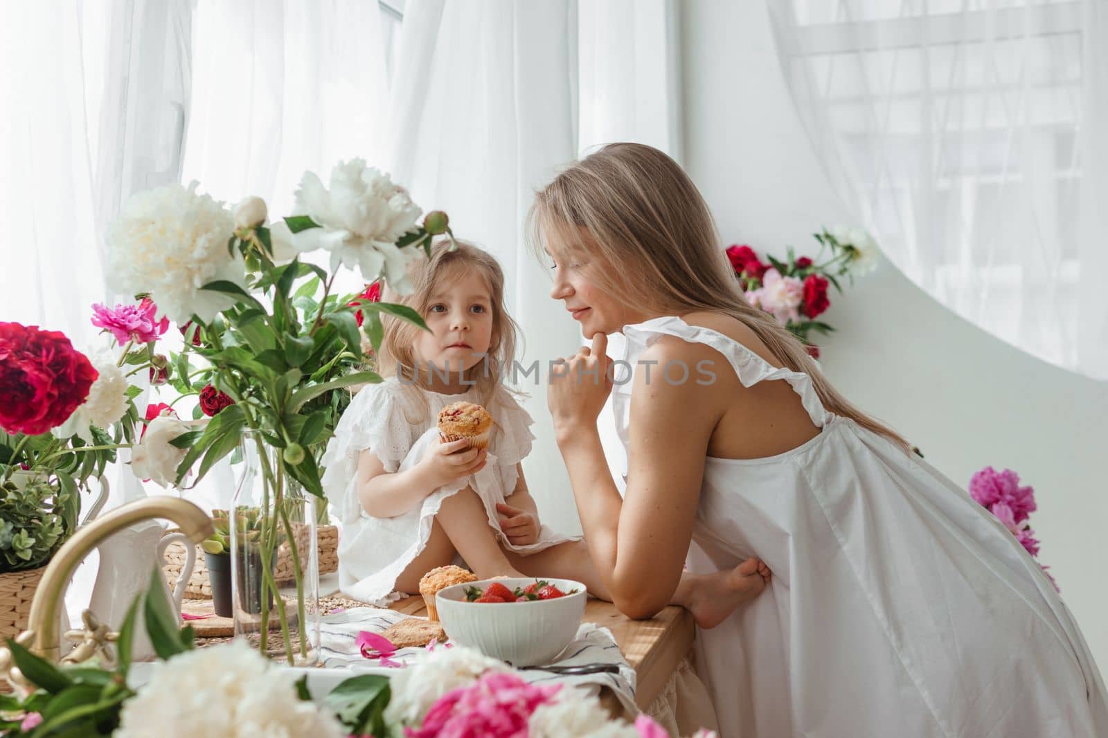 A little blonde girl with her mom on a kitchen countertop decorated with peonies. The concept of the relationship between mother and daughter. Spring atmosphere.