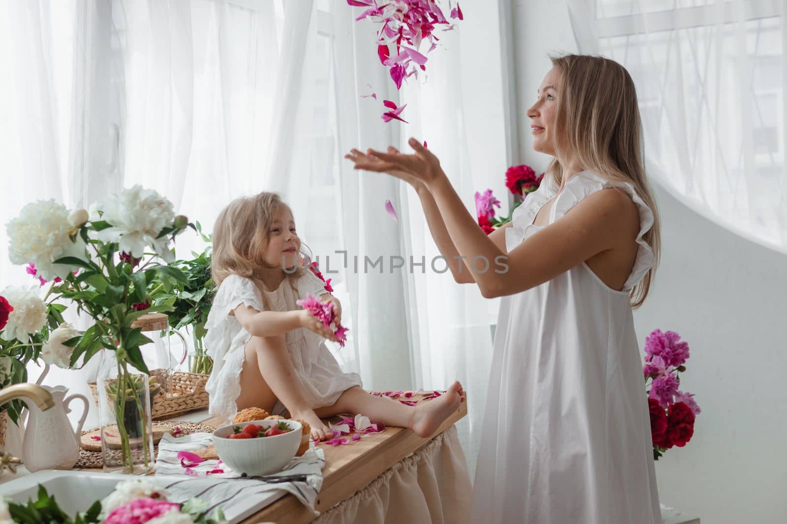 A little blonde girl with her mom on a kitchen countertop decorated with peonies. The concept of the relationship between mother and daughter. Spring atmosphere.