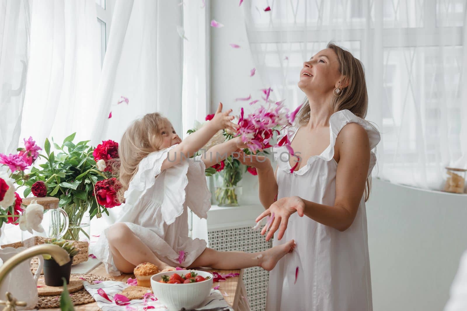 A little blonde girl with her mom on a kitchen countertop decorated with peonies. The concept of the relationship between mother and daughter. Spring atmosphere.