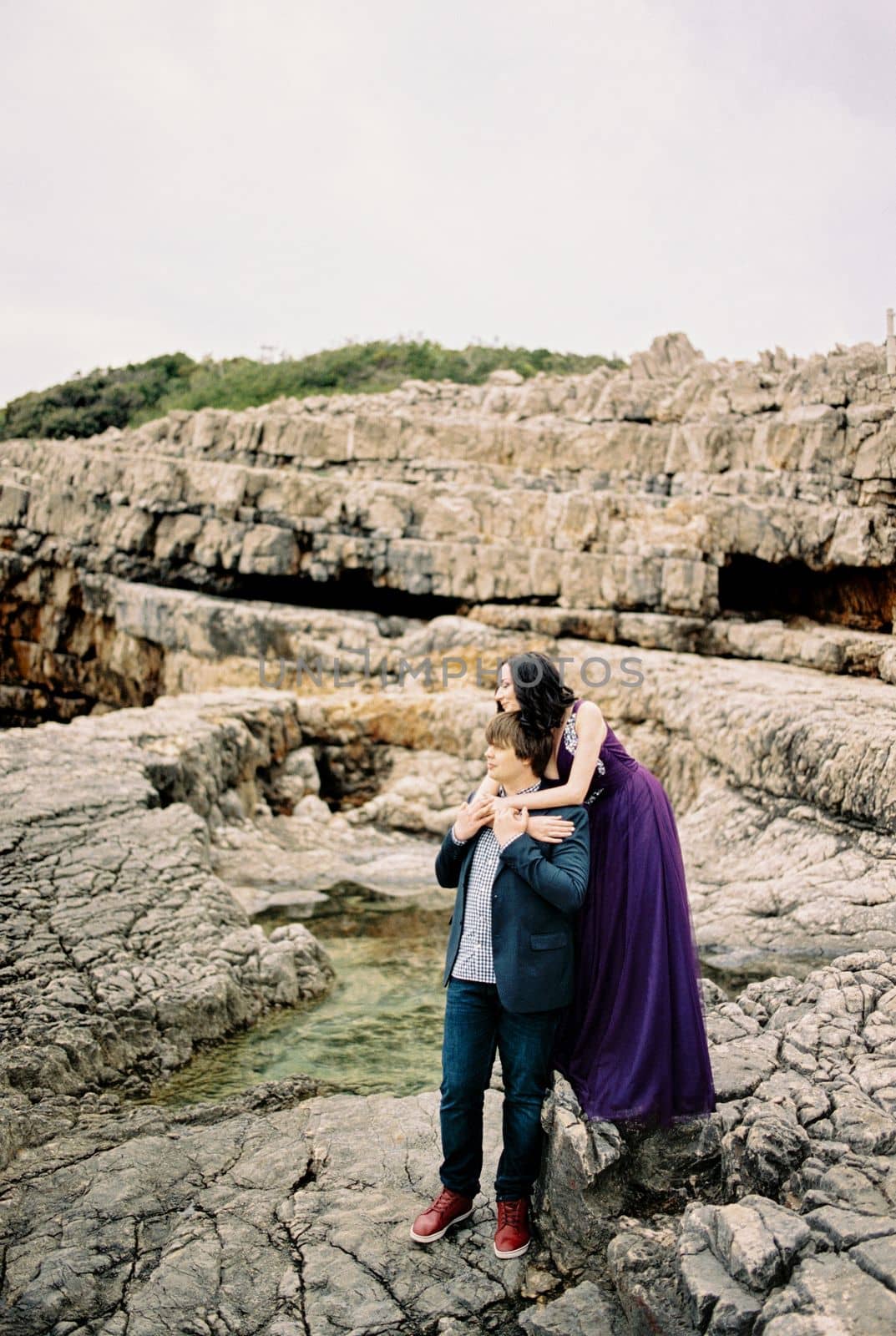 Woman in a lilac dress hugs man from behind standing on the rocks. High quality photo
