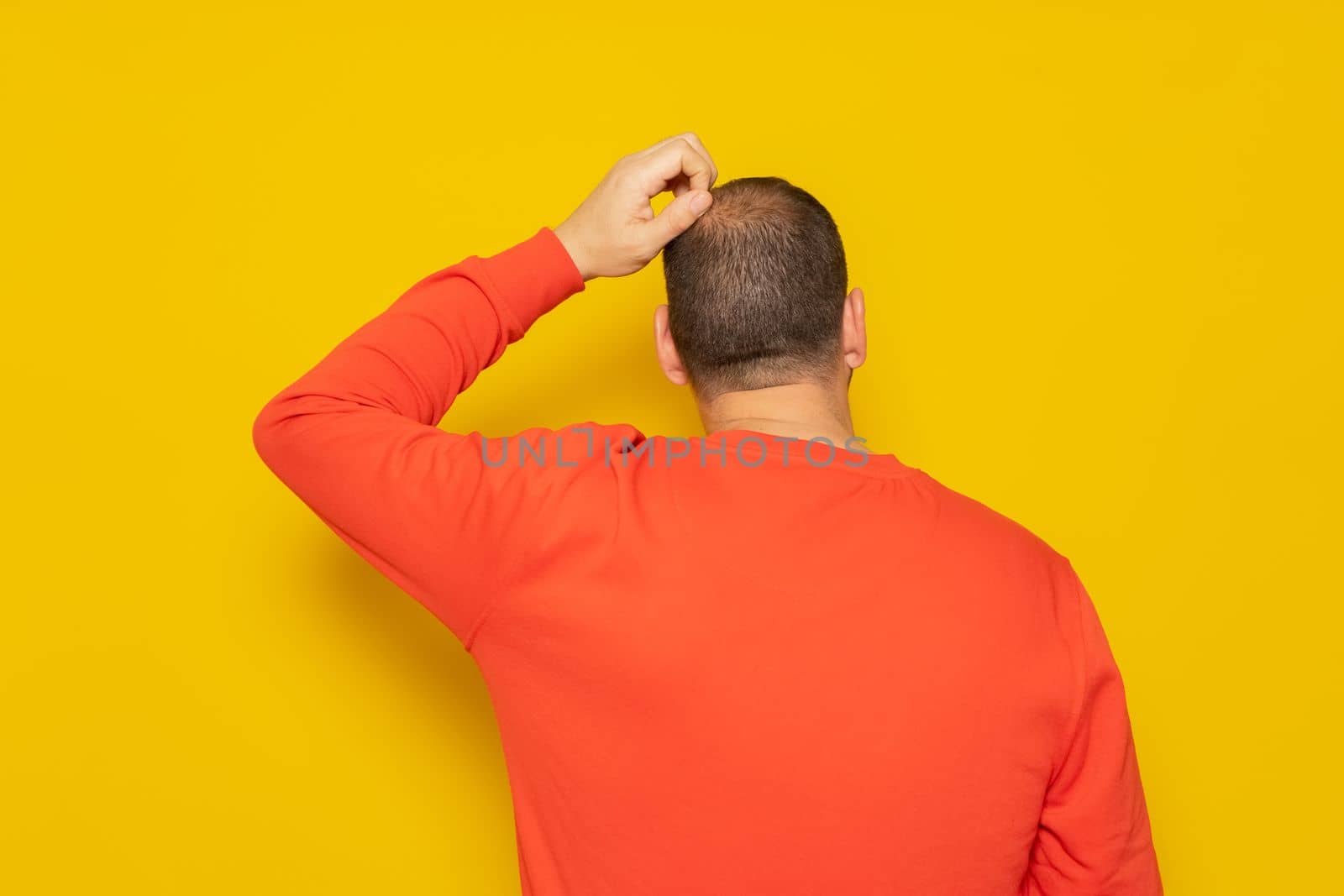 Back view of thoughtful man scratching his head over yellow background. He is doubting his existence, he does not understand the reason for life and what meaning it has