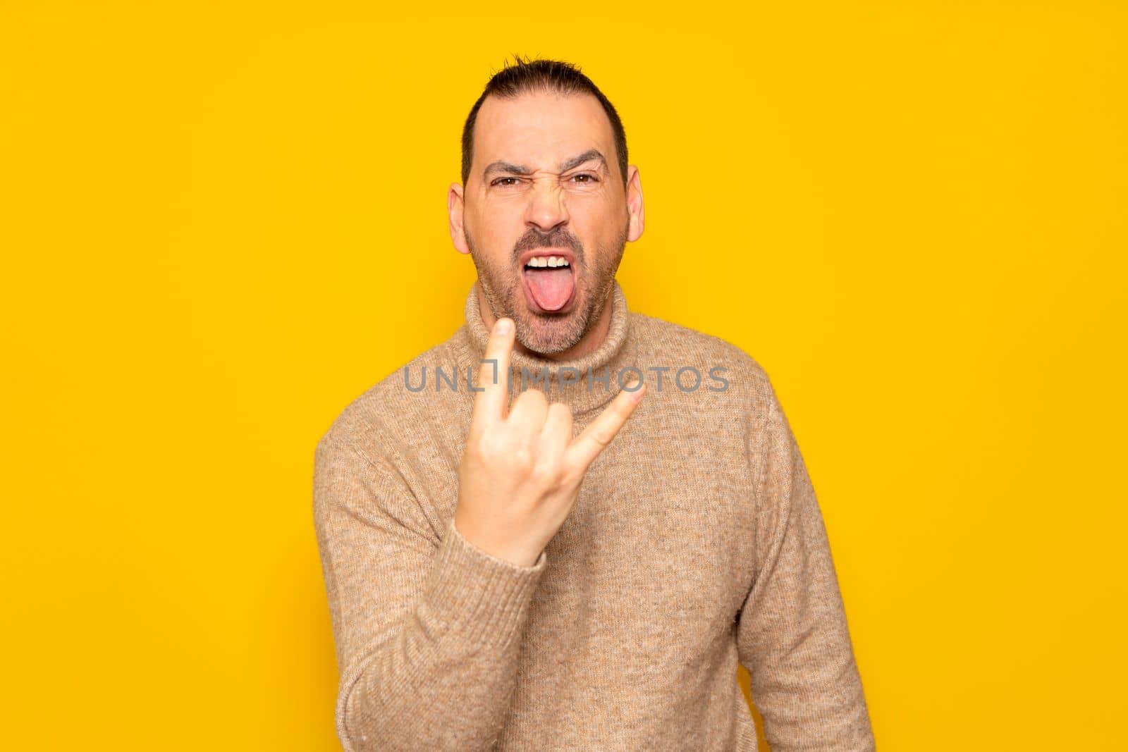 Bearded hispanic man wearing turtleneck isolated over yellow background showing rock gesture with fingers and sticking out tongue. Concept of rebellion and anarchism