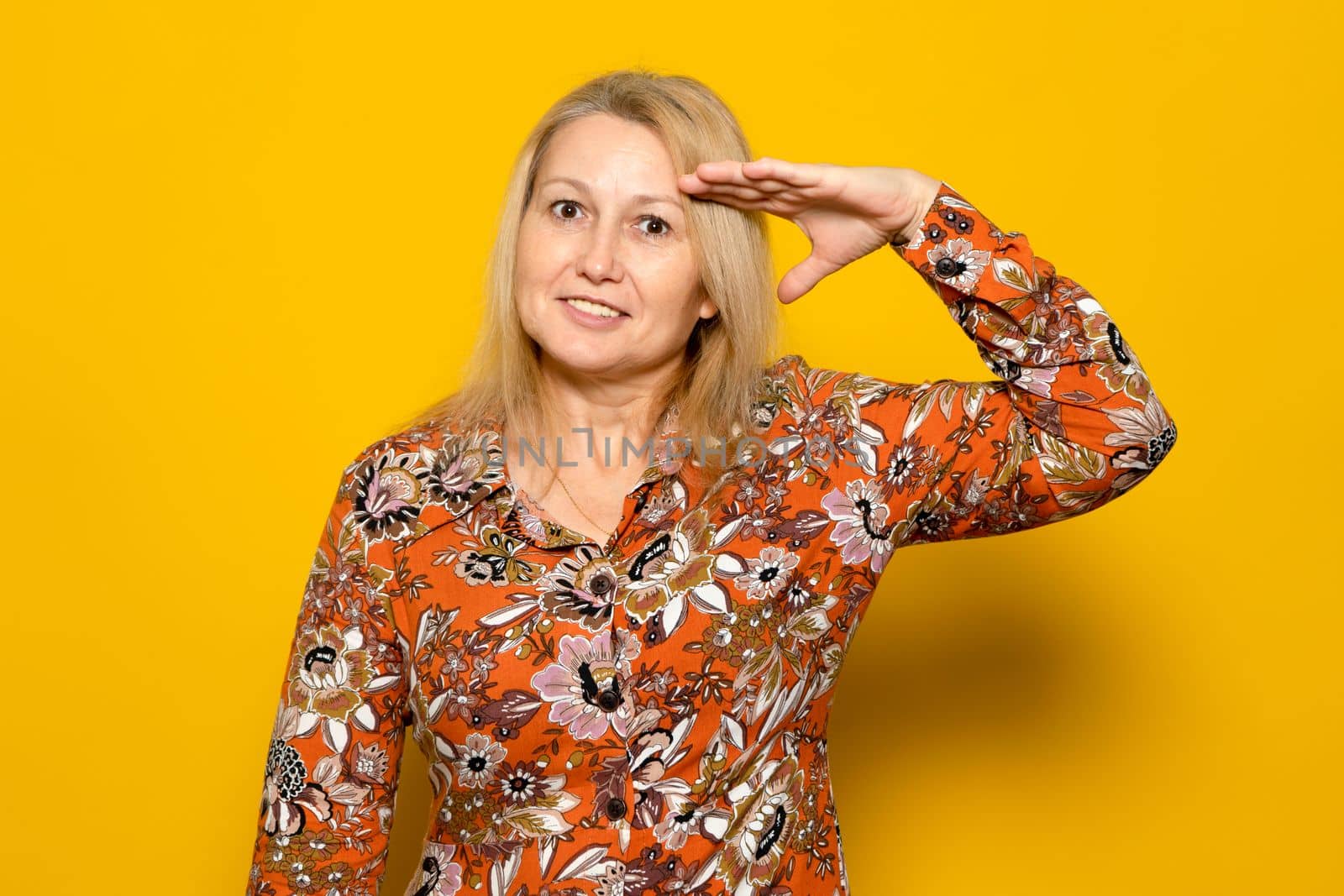 Caucasian blonde woman in a patterned dress doing the military salute isolated over yellow background, fulfilling the homeland