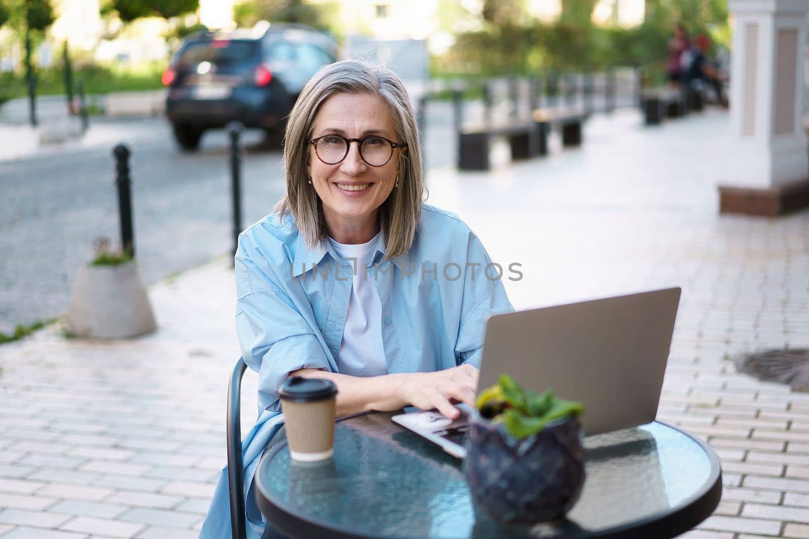 Happy and content Caucasian mature woman with long grey hair, seated at a street cafe with a notebook. She appears relaxed and enjoying her leisure time while staying connected with the world through the convenience of technology.