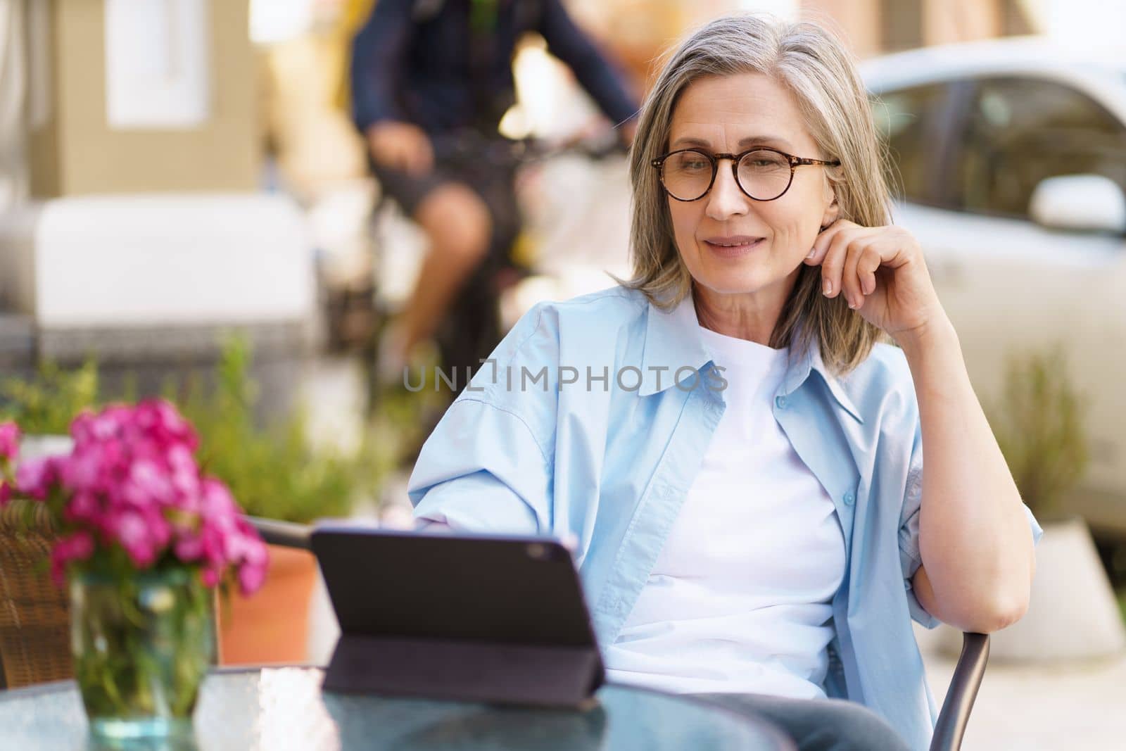 Mature European woman seated at an outdoor cafe, browsing information on her tablet PC. She appears relaxed and content, enjoying her leisure time while staying connected through the convenience of technology. High quality photo